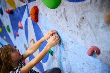 6 yaşında bir çocuk tırmanmaya odaklanmış, dikkatli bir şekilde kapalı bir kaya duvarına tutunuyor. Renkli ambarlar ve detaylı konsantrasyon kararlılığını gösteriyor..