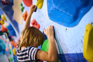 6 yaşında bir çocuk tırmanmaya odaklanmış, dikkatli bir şekilde kapalı bir kaya duvarına tutunuyor. Renkli ambarlar ve detaylı konsantrasyon kararlılığını gösteriyor..