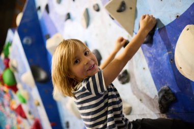 6 yaşında gülümseyen bir çocuk, renkli kolları olan bir kaya duvarına tırmanıyor ve kameraya bakıyor. Yüzündeki ifade, heyecanı ve kendine güveni gösteriyor..