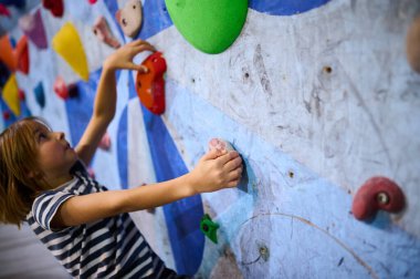 6 yaşında bir çocuk tırmanmaya odaklanmış, dikkatli bir şekilde kapalı bir kaya duvarına tutunuyor. Renkli ambarlar ve detaylı konsantrasyon kararlılığını gösteriyor..