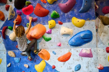 6 yaşında bir çocuk tırmanmaya odaklanmış, dikkatli bir şekilde kapalı bir kaya duvarına tutunuyor. Renkli ambarlar ve detaylı konsantrasyon kararlılığını gösteriyor..