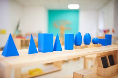 Montessori geometric solids in a preschool classroom. Bright blue shapes on wooden bases, including cubes, spheres, and cones, arranged on a wooden shelf. clipart