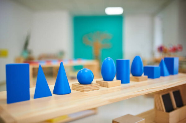 Montessori geometric solids in a preschool classroom. Bright blue shapes on wooden bases, including cubes, spheres, and cones, arranged on a wooden shelf.