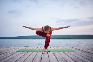 Sakin bir gölün yakınındaki ahşap iskelede yoga yapan bir kadın. Ardha Baddha Padmottanasana Yarı Zıplayan Lotus Yeşil yoga minderinin üstünde kırmızı bir elbise içinde duruyor.