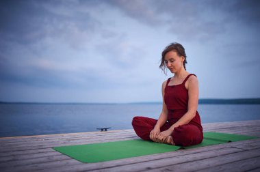 Sakin bir gölün kenarındaki ahşap iskelede yoga yapan bir kadın. Akşam boyunca yeşil yoga minderinde kırmızı kıyafetli Sukhasana Easy Poz veriyordu..