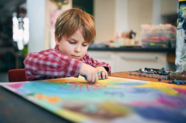 Ekose gömlekli genç bir çocuk renkli pasteller çiziyor. Evdeki büyük bir kağıt parçasına. Rahat bir sanat alanındaki yaratıcı aktivitelere odaklanmış..