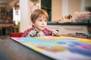 Ekose gömlekli genç bir çocuk renkli pasteller çiziyor. Evdeki büyük bir kağıt parçasına. Rahat bir sanat alanındaki yaratıcı aktivitelere odaklanmış..