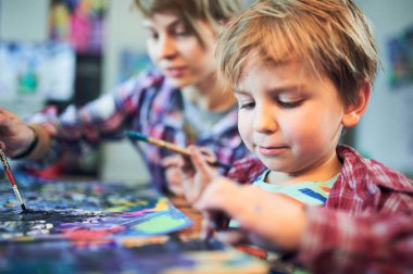 Genç bir çocuk fırçayla resim yaparken bir kadın ona bir sanat stüdyosunda ahşap bir masada yardım ediyor. Arkaplanda canlı sanat eserleri ve araçlar