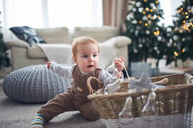 Süslü ağaçlar ve rahat mobilyalarla çevrili, neşeli bir atmosferin tadını çıkaran küçük bir çocuk, elinde cam Noel süsleri tutarak yumuşak bir halıya oturur..