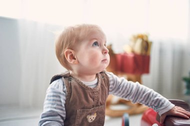 Küçük bir çocuk, kapalı bir alanda renkli oyuncaklarla meşgul olur ve merakla yukarı bakar. Yumuşak doğal ışık, neşeli atmosferi güçlendirir, sıcak bir his yaratır..