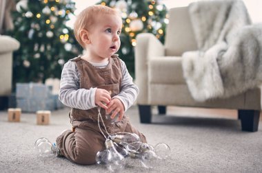 Genç bir çocuk, Noel ağacı ve sıcak bir oturma odasında yumuşak mobilyalarla çevrili bir dizi dekoratif ışık tutarak halıya oturur..