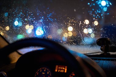 Arabanın ön camına yağmur damlaları damlıyor ve gece şehir ışıkları bulanık, içeriden görülüyor. Yumuşak ışıkla aydınlatılmış direksiyon ve gösterge paneli.