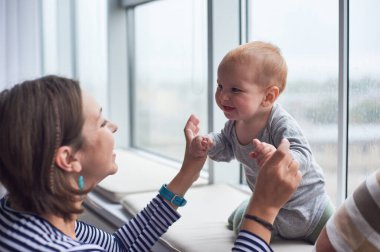 Neşeli bir bebek pencere kenarında oturur, gülümser ve annesiyle oynar. Yağmur, keyifli etkileşimlerini ve bağlantılarını geliştirmek için rahat bir zemin sağlar..