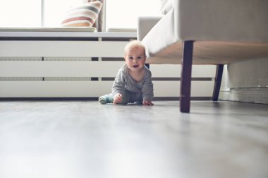 Bir bebek, parlak ve modern bir oturma odasında pürüzsüz bir ahşap zeminde sürünüyor. Doğal ışık geniş pencerelerden içeri akar ve sıcak bir atmosfer yaratır..