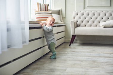 Bebek, rahat yastıklar ve yumuşak güneş ışığıyla dolu, parlak, modern bir oturma odasında, parlak bir pencere pervazına tutunurken güven içinde durur..