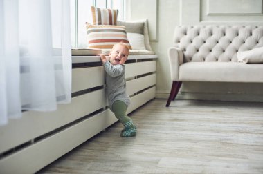 Küçük bir bebek, parlak bir oturma odasında beyaz bir radyatöre tutunurken mutlu bir şekilde gülümsüyor. Mekanda rahat bir kanepe ve dekoratif yastıklar var..