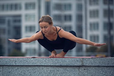Yetenekli bir yoga pratisyeni çatıda dengede durur, zorlu bir duruşta güç ve esneklik gösterir. Şehir manzarası çarpıcı bir zemin sunuyor, modern binalar sergileniyor..