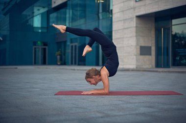Yetenekli bir yoga uygulayıcısı, gece boyunca modern bir şehir ortamında kırmızı bir paspasın üzerinde zorlu bir duruş sergiliyor. Parlak binalar bu odaklanmış faaliyet için sakin bir zemin oluşturur..