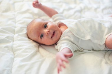 Bir bebek, yumuşak beyaz bir yatak örtüsünde kolları açık uzanarak mutlu bir şekilde yatar. Samimi ortam, parlak gözler, büyüleyici bir gülümseme masumiyet ve neşe saçıyor..