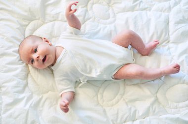 Beyaz tulumlu neşeli bir bebek, aydınlık bir odadaki yumuşak bir yorganın üzerinde uzanıyor, gülümsüyor ve kollarını ve bacaklarını oynatıyor, neşeli ve huzurlu bir anın tadını çıkarıyor..