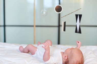 Newborn baby lying on a soft bed, reaching for a black and white Montessori Munari mobile, promoting early visual development and sensory engagement in infants. clipart