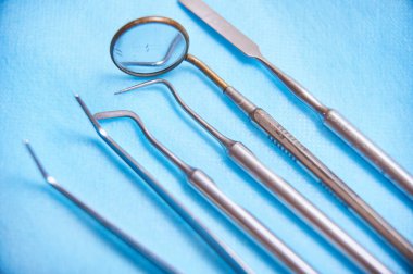 Muayene ve temizlik için kullanılan çeşitli diş aletleri mavi arka planda düzgün bir şekilde düzenlenmiştir. Kurulum, diş bakımında gerekli hassasiyeti vurguluyor.