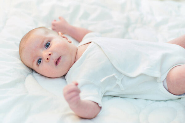 Маленький ребенок, одетый в простой белый наряд, лежит на мягком белом одеяле, глядя вверх с широкими, любознательными голубыми глазами. Солнечный свет наполняет яркую комнату, усиливая мирную атмосферу.