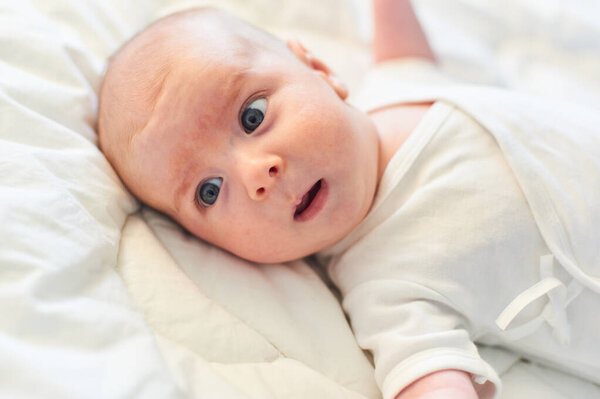 A baby with bright blue eyes lies on a fluffy white blanket, displaying an inquisitive expression. The soft lighting highlights the gentle features, creating a serene atmosphere.