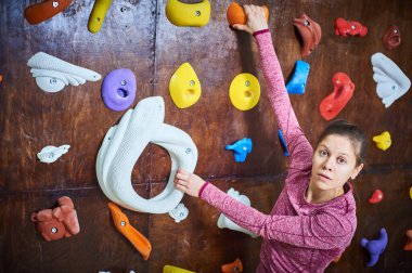 Bir dağcı, çeşitli renkli tutacaklarla kaplı kapalı bir tırmanma duvarına uzanırken dikkatle odaklanır. Burası modern bir tırmanma salonu..