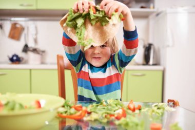 Gülümseyen çocuk mutfakta sebzelerle oynuyor ve tortilla dürümü, renkli çizgili gömlek, dağınık masa, eğlence ve neşe dolu an, sağlıklı yemek hazırlığı.