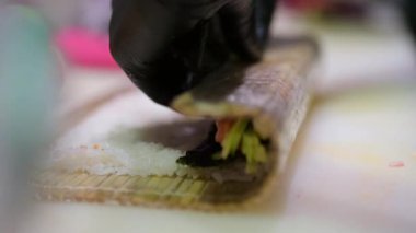 Eldivenli suşi, pilav, somon, salatalık ve bambu hasırı üzerinde nori, profesyonel bir mutfak ortamında yemek hazırlama sürecinin yakın çekimi..