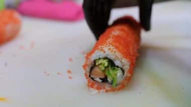 Somon, salatalık ve krem peynirle kaplı turuncu tobiko ile kaplı suşi rulosu beyaz bir yüzeyde dilimlenerek hazırlanma sürecinin yakınına getiriliyor..