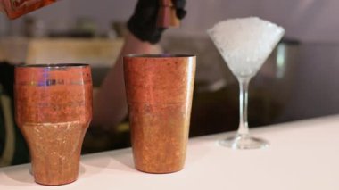 Barmen jigger kullanarak bakır bir kokteyl bardağına sıvı dolduruyor. Arka planda buzla dolu bir martini bardağı var..
