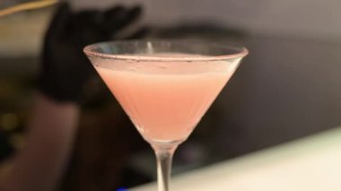 Barmen, martini bardağında pembe bir kokteyl süslüyor. Siyah eldivenler giyiyor. İçki sunumunda profesyonelliği ve zarafeti vurguluyor..