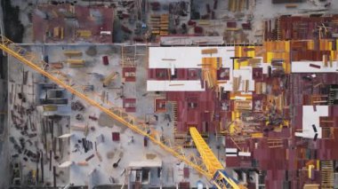 İnşaat işçileri büyük bir sitede çeşitli malzeme ve ekipmanları istiflemekle ve düzenlemekle meşguller. Üstümüzde sarı bir vinç kulesi var, önemli bir bina faaliyeti gösteriyor..