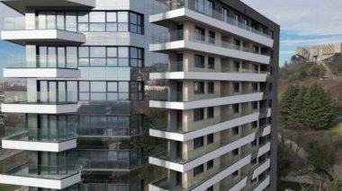 Çağdaş konut binasında birden fazla balkonu olan çarpıcı bir cam cephe sergileniyor. Çevresi yeşilliklerle çevrili yapı, açık bir gökyüzünün altında hareketli bir şehir ortamında duruyor..