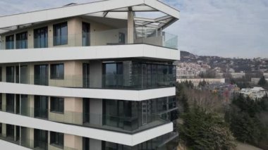 Şehir manzarasında yeşilliklerle çevrili geniş balkonların bulunduğu çağdaş, çok katlı bir apartman. Tasarım, geniş pencereli modern mimariyi vurguluyor.
