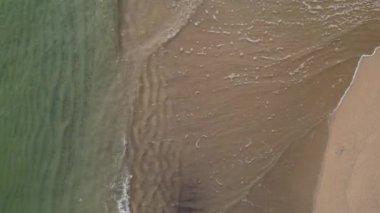 Kıyı şeridinde turkuaz dalgalar altın kumu karşılayarak çarpıcı bir kontrast oluşturuyor. Hafif dalgalar güneş ışığını yansıtıyor, sakin bir sahil atmosferi sergiliyor..