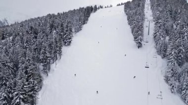Kayakçılar buzla kaplı ağaçlarla çevrili karlı bir yamaçta süzülüyorlar. Bir kayak asansörü arka planda yükselirken gri bulutlar gökyüzünü kaplıyor ve kış atmosferi yaratıyor. Bansko, Bulgaristan