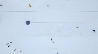 Kayakçılar ve snowboardcular güneşli bir günde beyaz manzaraya zıt parlak ceketlerle bir kayak asansörünün altındaki karlı arazide gezinirler..