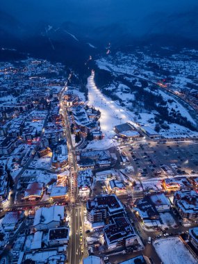 Alacakaranlıkta, karla kaplı yollar, hareketli dükkanlar ve manzarayı süsleyen parıldayan ışıklarla pitoresk bir kış köyü parıldıyor. Sakin dağlar manzarayı tamamlar..