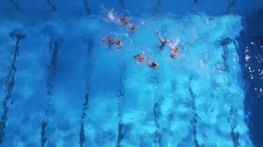 Mavi havuzda senkronize yüzücülerin performanslarının yukarıdan aşağı görüntüsü. Dinamik hareket ve sıçramalar, spor, takım çalışması ve su teması için ideal enerjik ve sanatsal bir sahne yaratır..