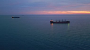 Illuminated cargo ship bulk carrier anchored on the calm sea at twilight, with city lights visible on the horizon.