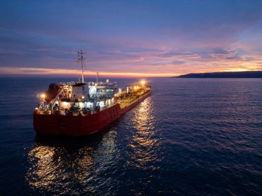 Alacakaranlıkta açık deniz ve ışıldayan ufukla çevrili parlak bir tanker gemisinin havadan görüntüsü..