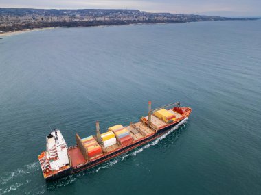 Bulgaristan 'ın Varna limanına giren bir kargo gemisinin hava görüntüsü. Gemi, Karadeniz 'in bulutlu sularında sanayi binaları, vinçler ve sakin sularıyla çevrili renkli konteynırlarla dolu.