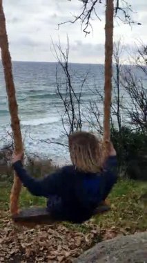 Okyanus manzaralı, kırsal bir ipte sallanan bir çocuk. Ağaçların ve dalgaların huzurlu çevresi doğanın ve çocukluğun basitliğini vurgulayan neşeli ve huzurlu bir an yaratıyor.