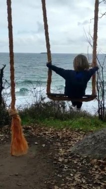 Okyanus manzaralı, kırsal bir ipte sallanan bir çocuk. Ağaçların ve dalgaların huzurlu çevresi doğanın ve çocukluğun basitliğini vurgulayan neşeli ve huzurlu bir an yaratıyor.