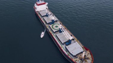 Açık denizde kızıl bir pruvası olan büyük bir kargo gemisi, bulutlu bir gökyüzünün altında bir römorkör eşliğinde, deniz taşımacılığı sahnesi..
