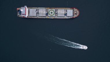 Açık denizde kızıl bir pruvası olan büyük bir kargo gemisi, bulutlu bir gökyüzünün altında bir römorkör eşliğinde, deniz taşımacılığı sahnesi..
