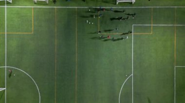 Yapay ışıklar altında, oyuncuların ve ekipmanların görülebildiği yeşil bir sahada futbol antrenmanının yukarıdan aşağı görüntüsü.